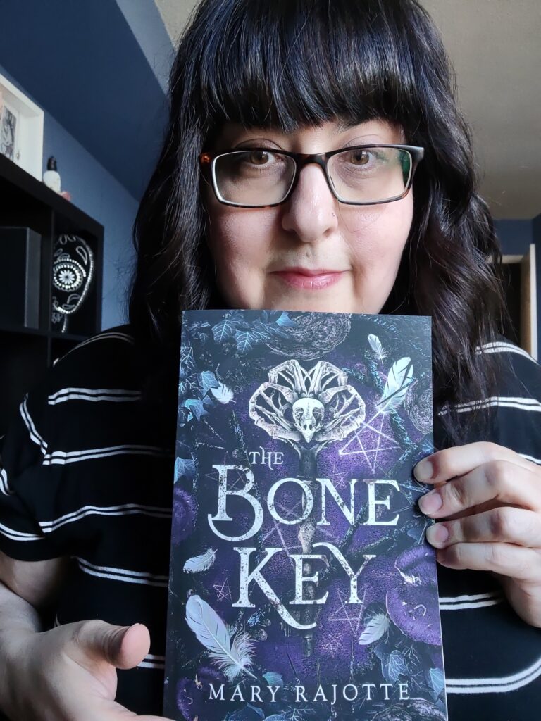 A photo of Mary Rajotte holding her paperback boo, The Bone Key. The cover has an aged key made from a bone and owl skull in the middle.