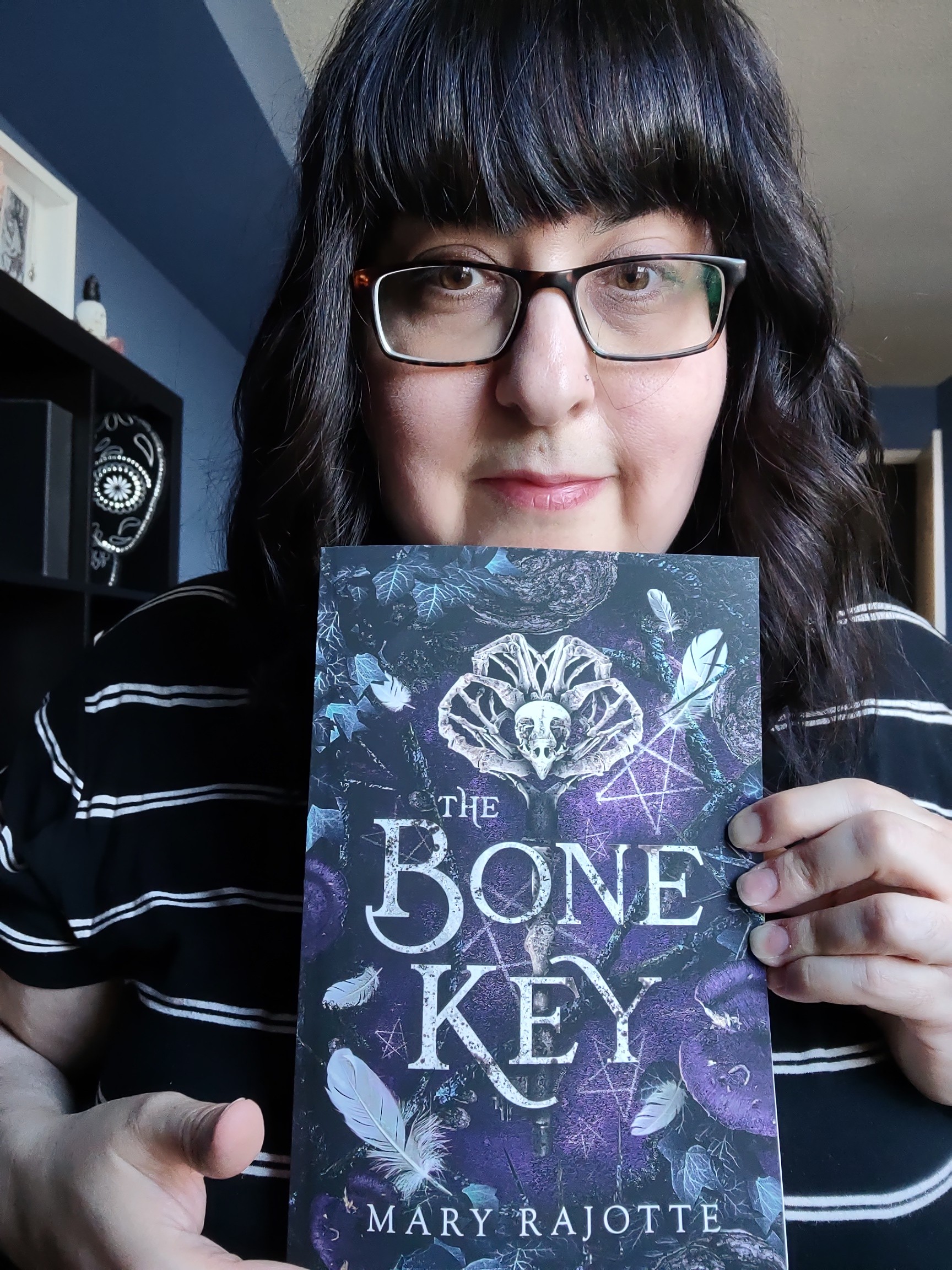 A photo of Mary Rajotte holding her paperback boo, The Bone Key. The cover has an aged key made from a bone and owl skull in the middle.