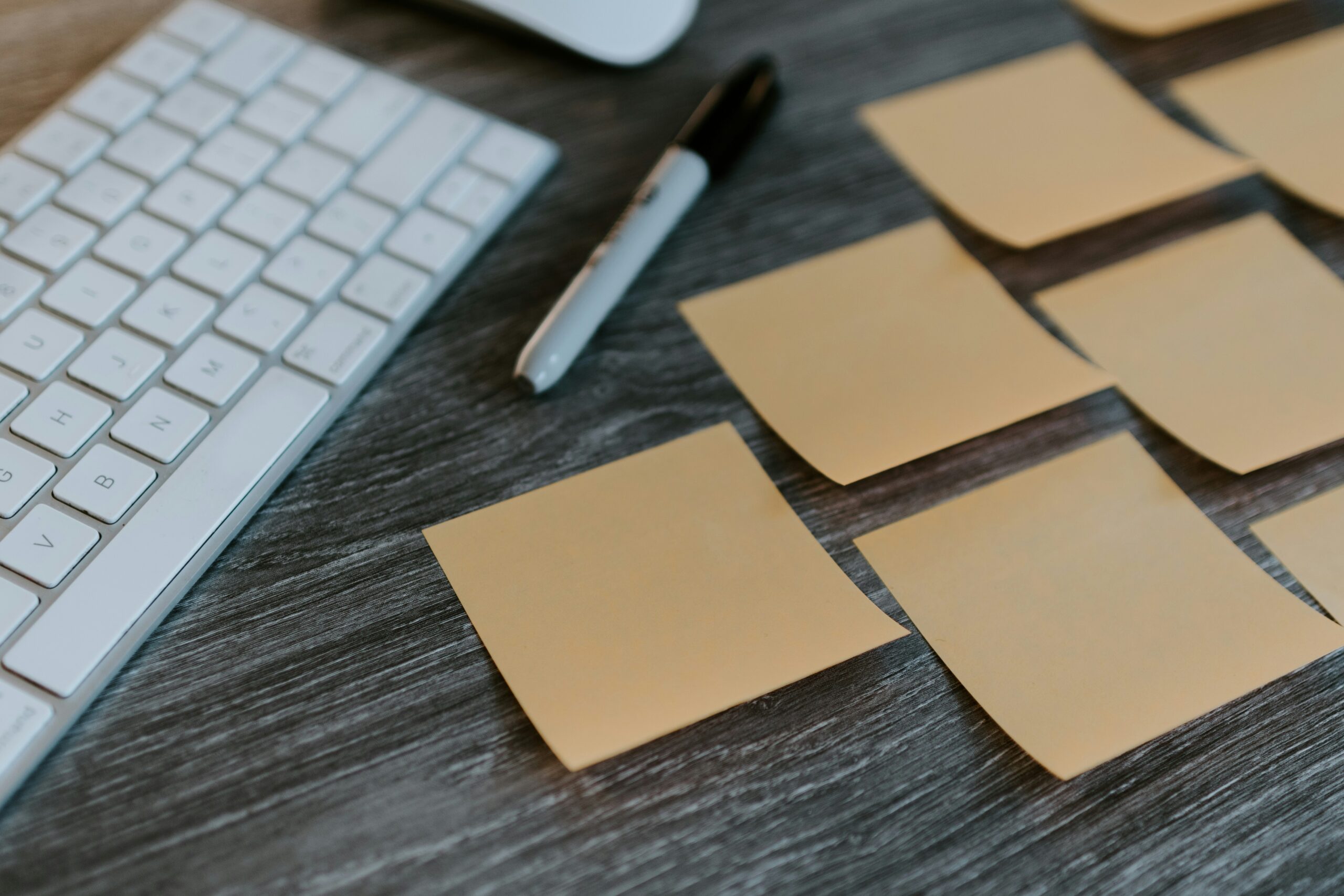 A close-up image of a dark grey desk with a white computer keyboard on the left side. Beside the keyboard there are 7 blank kraft paper sticky note and a black sharpie pen